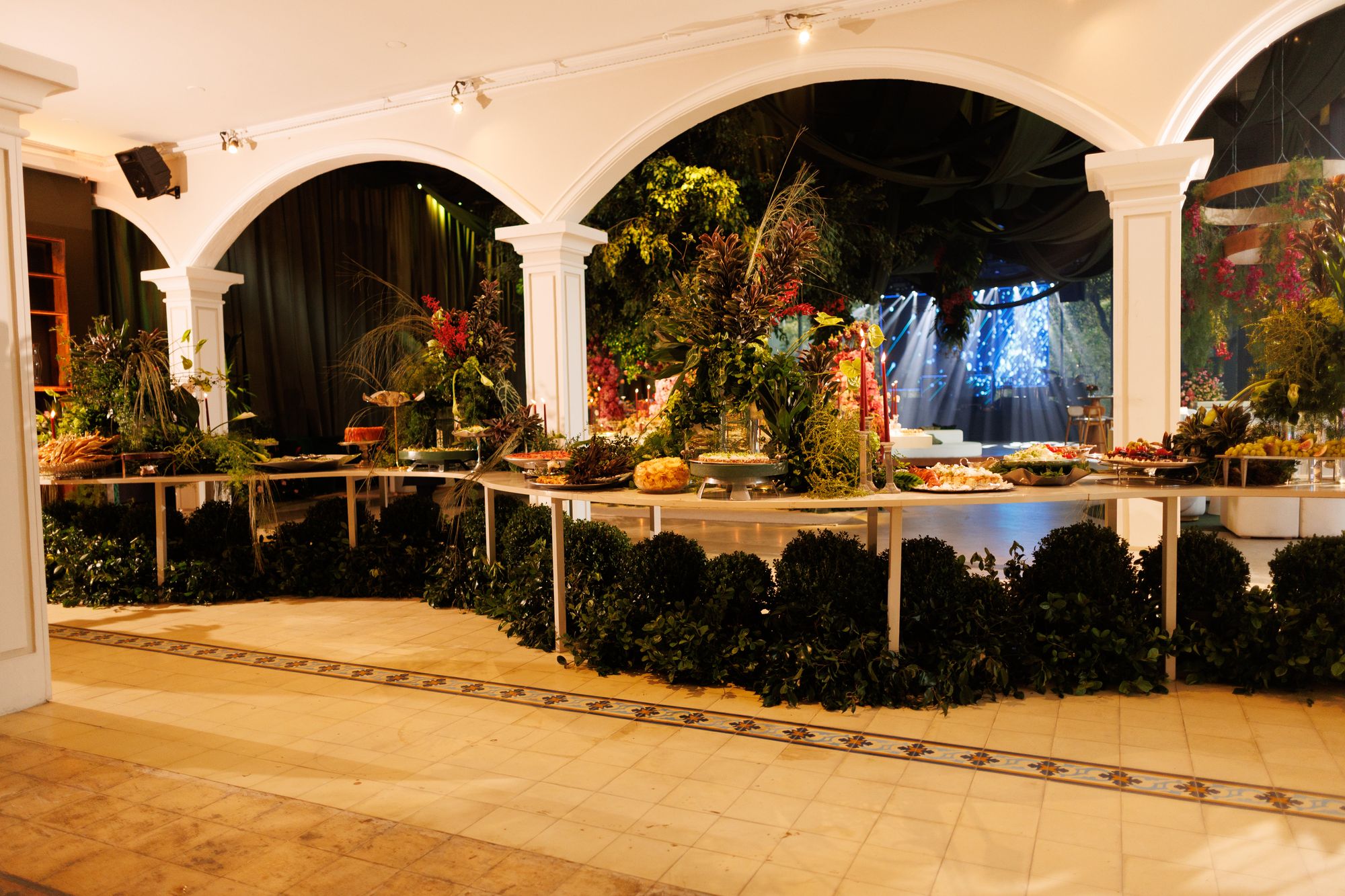 Buffet montado sob os arcos do salão, com arranjos de plantas, velas e pratos servidos